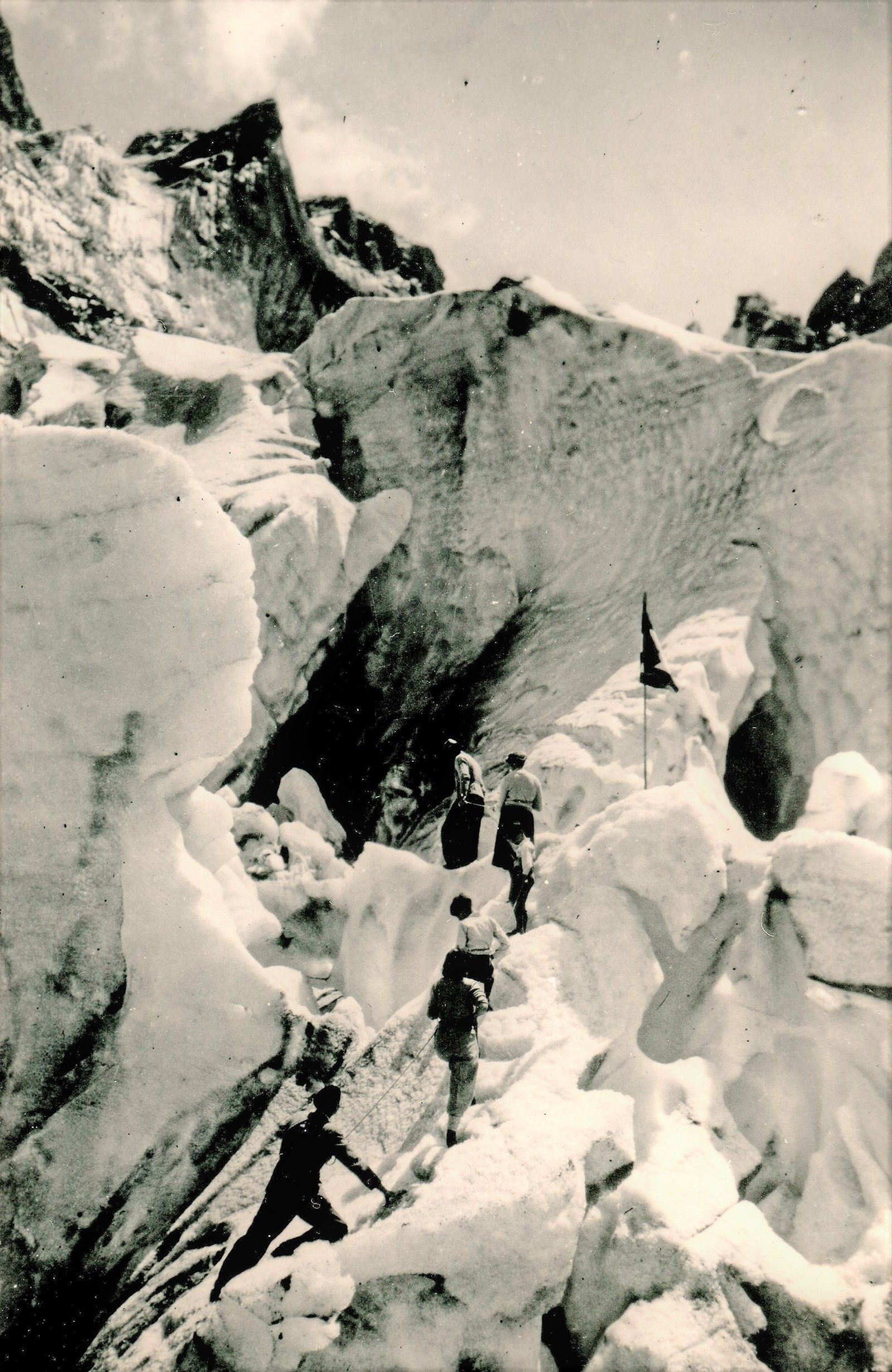 A black and white photograph showing a group of six people walking up the steep sides of a glacier, tethered together by rope.