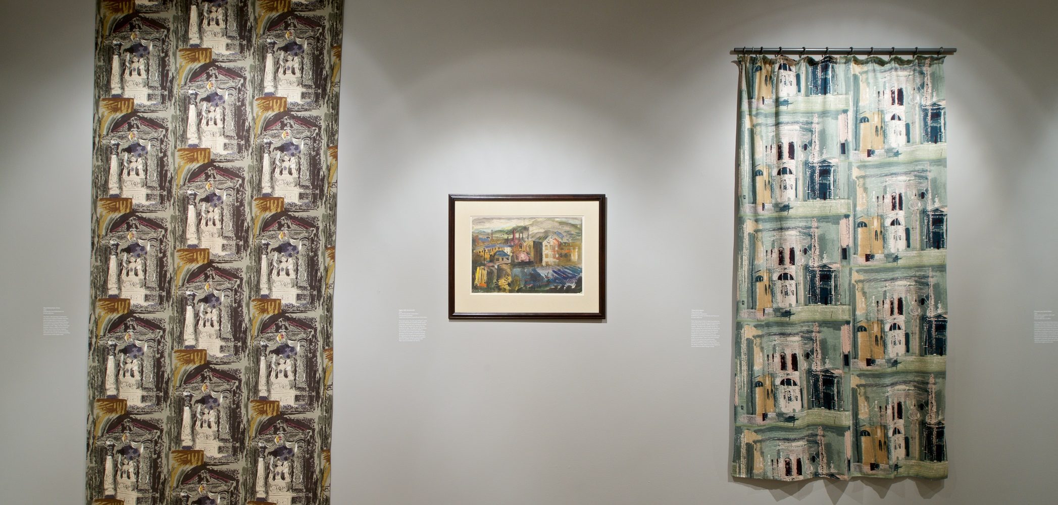 View of interior of gallery showing two lengths of cloth with two different abstract designs. Between them is a painting showing a mid-century urban landscape.