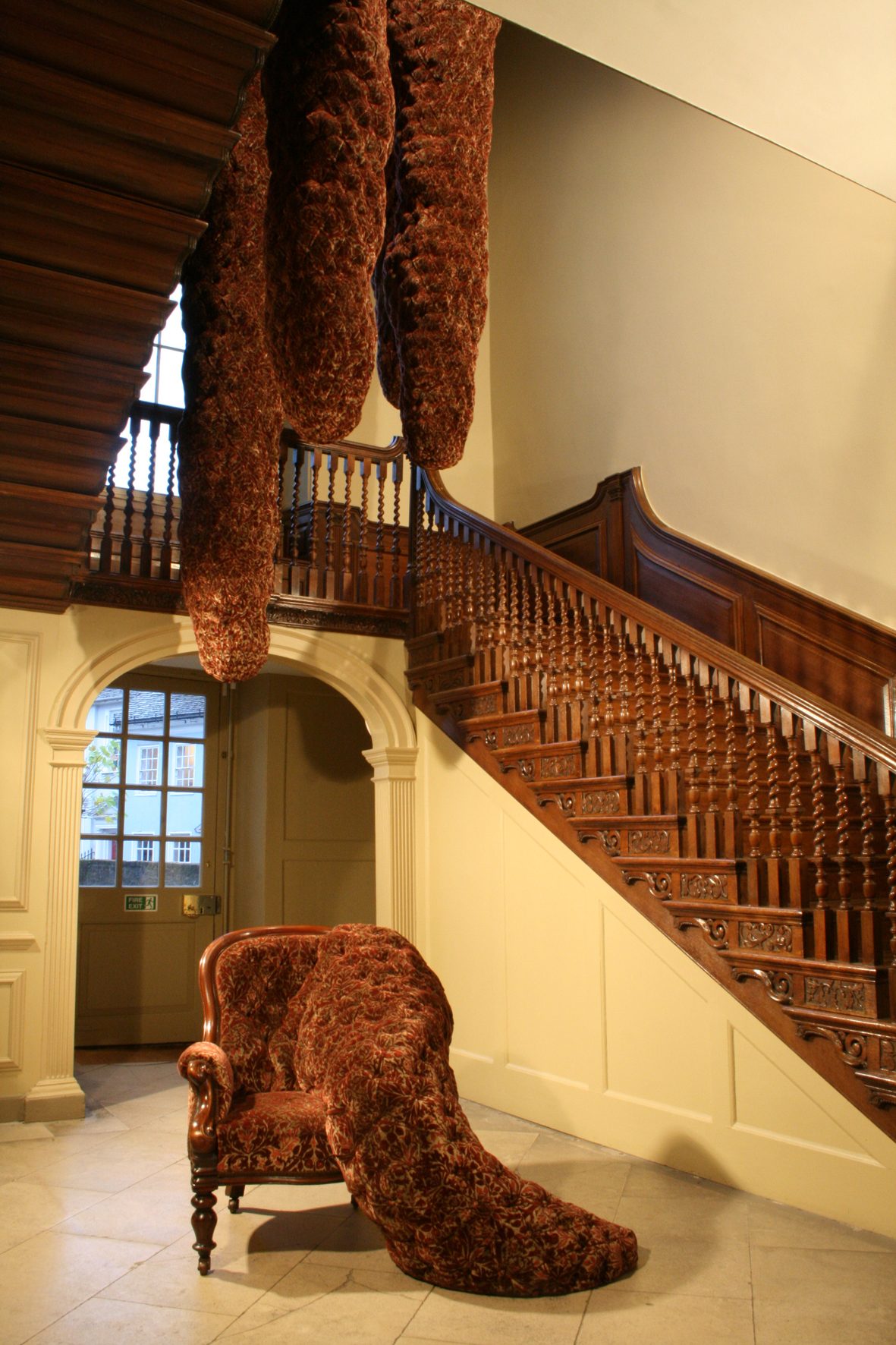 A period staircase with a red upholstered armchair with amorphous bulging out of one side at the foot of the staircase. More long upholstered forms hang down the centre of the stairwell.